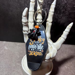 May include: A black motel-style keyring shaped like a coffin with a small black ghost figure wearing an orange pumpkin hat on top. The keyring features the white and orange text "Spooky is my favorite season" and includes a silver metal ring.