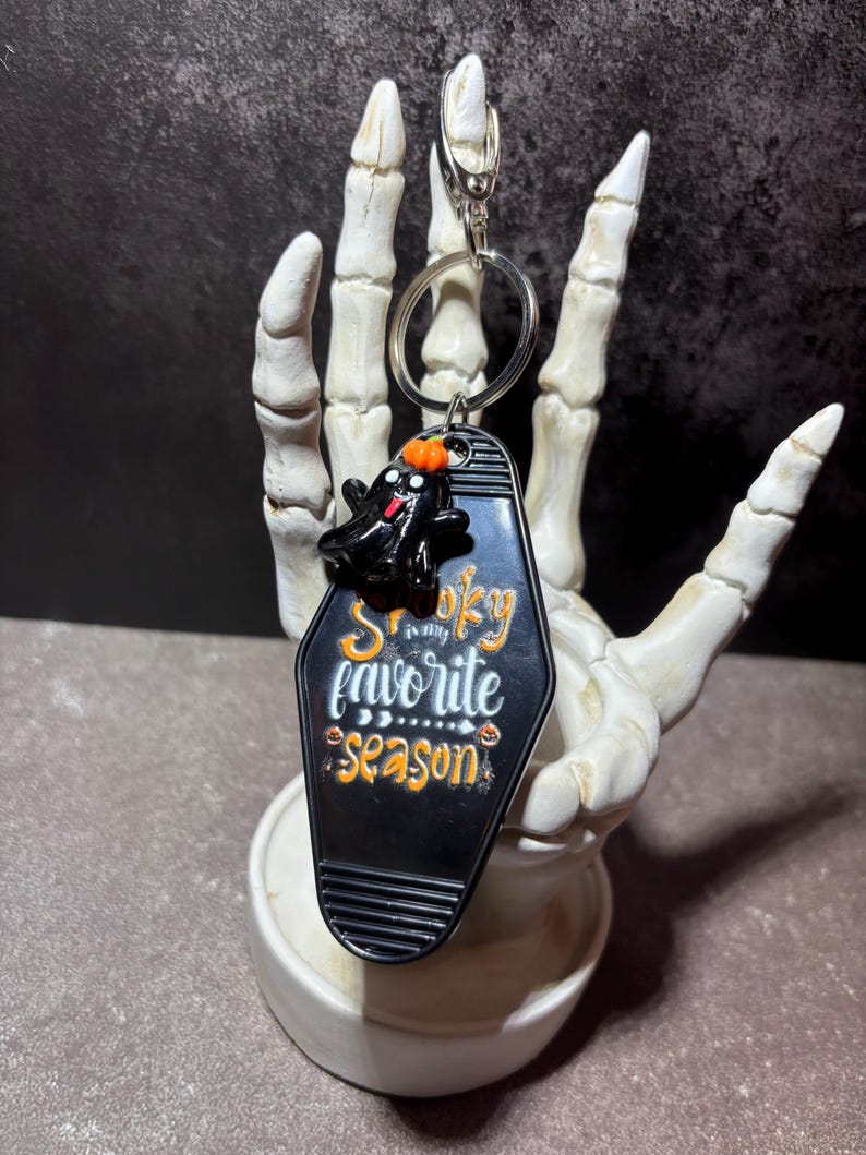 May include: A black motel-style keyring shaped like a coffin with a small black ghost figure wearing an orange pumpkin hat on top. The keyring features the white and orange text "Spooky is my favorite season" and includes a silver metal ring.