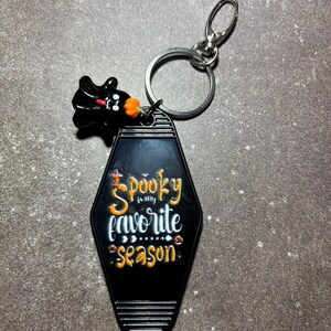 May include: A black motel-style keyring with a silver metal ring and lobster clasp. The keyring features the orange and white text "Spooky is my favourite season" and a small black ghost charm with an orange pumpkin accent attached to the ring.