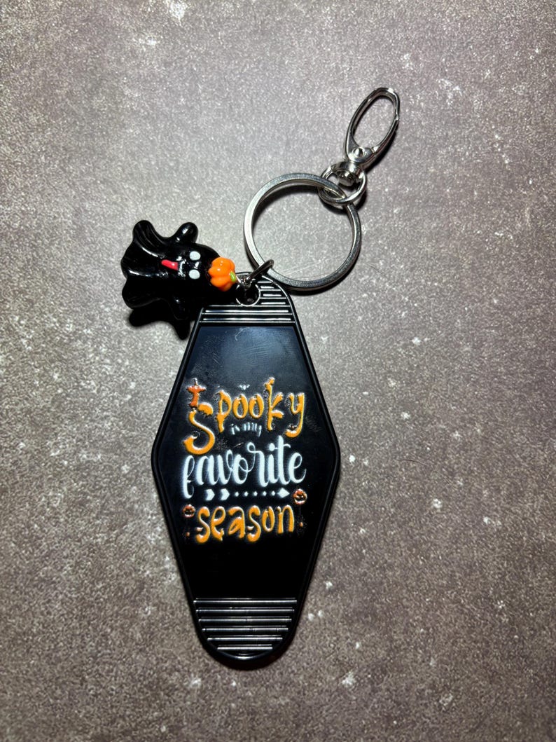 May include: A black motel-style keyring with a silver metal ring and lobster clasp. The keyring features the orange and white text "Spooky is my favourite season" and a small black ghost charm with an orange pumpkin accent attached to the ring.