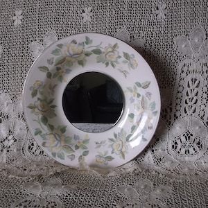 May include: A white ceramic plate with a floral design and a round mirror in the center. The plate has a gold rim and is decorated with yellow, green, and blue flowers.