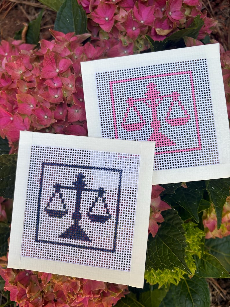 Puede incluir: Dos lienzos cuadrados de punto de aguja con un dise&ntilde;o de balanza de la justicia. Un lienzo muestra la balanza en hilo rosa vibrante, mientras que el otro lienzo presenta el mismo dise&ntilde;o de balanza bordado en hilo azul marino oscuro sobre malla blanca.