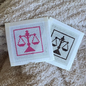 Puede incluir: Dos lienzos cuadrados de punto de aguja con un dise&ntilde;o de balanza de la justicia. Un lienzo muestra la balanza en hilo rosa brillante, mientras que el otro presenta el mismo dise&ntilde;o en hilo negro. Ambos lienzos tienen un borde de malla blanca.