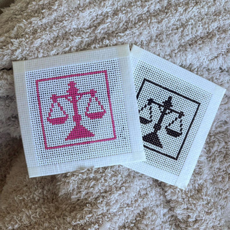Puede incluir: Dos lienzos cuadrados de punto de aguja con un dise&ntilde;o de balanza de la justicia. Un lienzo muestra la balanza en hilo rosa brillante, mientras que el otro presenta el mismo dise&ntilde;o en hilo negro. Ambos lienzos tienen un borde de malla blanca.