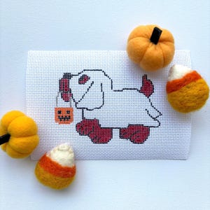 May include: A white cross-stitch dog holding a jack-o'-lantern bucket, surrounded by felt pumpkins and candy corn. The dog has red paws and a red tongue. The pumpkins are orange with black stems, and the candy corn is white, orange, and yellow.
