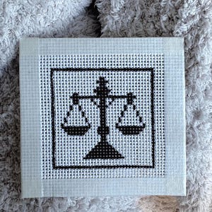 Puede incluir: Una pieza cuadrada de lienzo blanco para bordar con un dise&ntilde;o en punto de cruz negro de una balanza de la justicia. El dise&ntilde;o est&aacute; centrado dentro de un borde cuadrado negro y el lienzo tiene una textura de rejilla visible en toda la superficie.