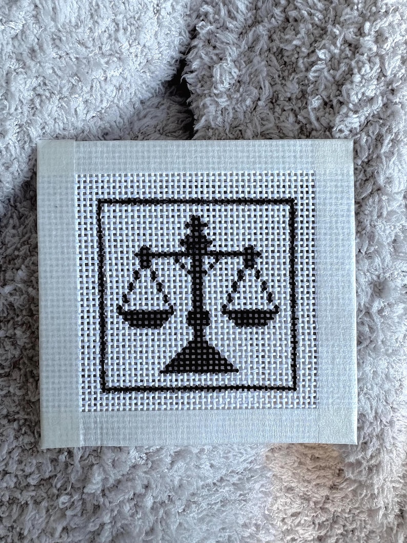 Puede incluir: Una pieza cuadrada de lienzo blanco para bordar con un dise&ntilde;o en punto de cruz negro de una balanza de la justicia. El dise&ntilde;o est&aacute; centrado dentro de un borde cuadrado negro y el lienzo tiene una textura de rejilla visible en toda la superficie.