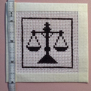 Puede incluir: Un trozo cuadrado de lienzo blanco para bordar con un dise&ntilde;o de punto de cruz marr&oacute;n oscuro de una balanza de la justicia centrada dentro de un borde cuadrado. Una cinta m&eacute;trica blanca est&aacute; colocada en el borde izquierdo, mostrando 10 cent&iacute;metros.