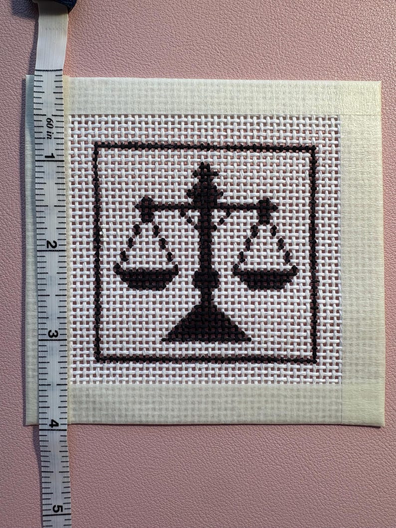 Puede incluir: Un trozo cuadrado de lienzo blanco para bordar con un dise&ntilde;o de punto de cruz marr&oacute;n oscuro de una balanza de la justicia centrada dentro de un borde cuadrado. Una cinta m&eacute;trica blanca est&aacute; colocada en el borde izquierdo, mostrando 10 cent&iacute;metros.