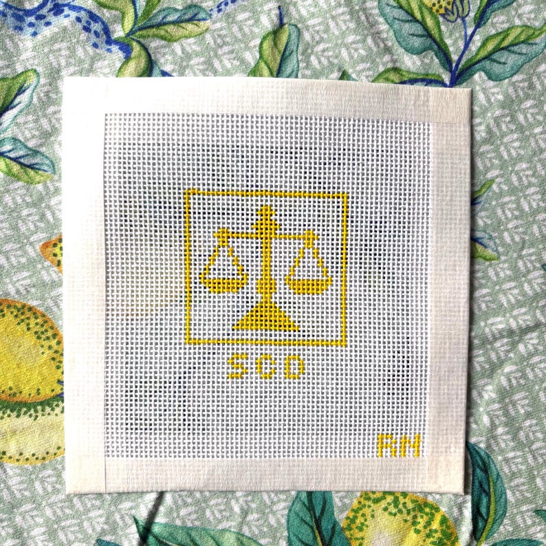 Puede incluir: Un lienzo cuadrado blanco para punto de aguja con un dise&ntilde;o amarillo bordado de una balanza de la justicia centrada dentro de un borde cuadrado. Debajo de la balanza, las letras SCD est&aacute;n bordadas en amarillo, con las letras RM en la esquina.