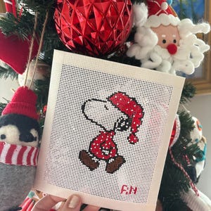 May include: A cross-stitched Snoopy Christmas ornament, wearing a red and white patterned outfit and hat. The ornament is held in front of a decorated Christmas tree with red ornaments and other festive decorations. The initials "R.N." are stitched on the ornament.