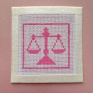 Puede incluir: Un lienzo cuadrado de punto de aguja con un icono de balanza de la justicia bordado en rosa brillante, centrado dentro de un borde cuadrado rosa. La tela de malla blanca est&aacute; enmarcada por una cinta de tela color crema claro en los cuatro lados.