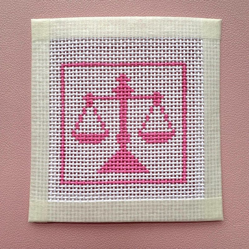 Puede incluir: Un lienzo cuadrado de punto de aguja con un icono de balanza de la justicia bordado en rosa brillante, centrado dentro de un borde cuadrado rosa. La tela de malla blanca est&aacute; enmarcada por una cinta de tela color crema claro en los cuatro lados.