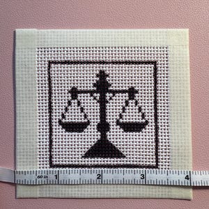 Puede incluir: Un trozo cuadrado de tela Aida blanca con un dise&ntilde;o de punto de cruz marr&oacute;n oscuro de una balanza de la justicia centrada dentro de un borde cuadrado. Una cinta m&eacute;trica blanca est&aacute; colocada a lo largo del borde inferior, indicando 10 cm.