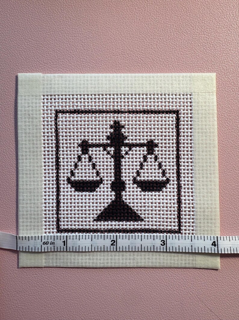 Puede incluir: Un trozo cuadrado de tela Aida blanca con un dise&ntilde;o de punto de cruz marr&oacute;n oscuro de una balanza de la justicia centrada dentro de un borde cuadrado. Una cinta m&eacute;trica blanca est&aacute; colocada a lo largo del borde inferior, indicando 10 cm.
