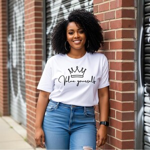 Puede incluir: Camiseta blanca con un gráfico de corona negra y el texto escrito "Value yourself". La camiseta se combina con unos vaqueros azules. La persona está apoyada en una pared de ladrillos rojos.