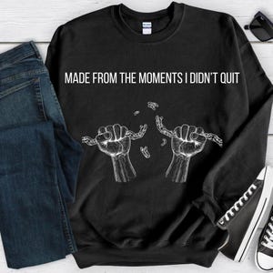 Puede incluir: Sudadera negra con el texto "MADE FROM THE MOMENTS I DIDN'T QUIT" en blanco. El diseño presenta dos manos rompiendo una cadena. Se ven unos vaqueros azules y zapatillas negras.