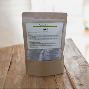 Può includere: Un sacchetto di carta marrone contenente bustine di tè alle foglie di lampone Passion Embrace. L'etichetta recita "Naturalmente senza caffeina | Supporto per il benessere femminile | 100% cotone". Il sacchetto contiene 40 bustine di tè.