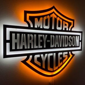 以下が含まれることがあります： 黒とオレンジ色のハーレーダビッドソンサイン。白い文字で「MOTOR HARLEY-DAVIDSON CYCLES」と書かれています。このサインはLEDライトで照らされており、精密にカットされたスチール製です。