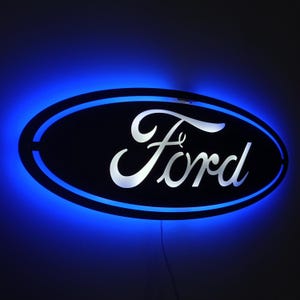 May include: A black oval sign with the word "Ford" in white, illuminated by a blue neon light. The sign has a blue outline and a wire extending from the bottom.