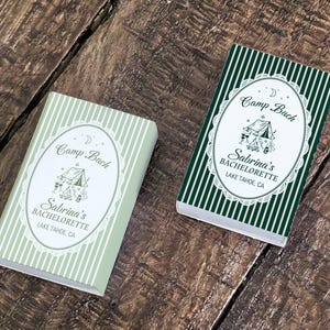 May include: Two rectangular boxes with the text "Camp Bach Sabrina's Bachelorette Lake Tahoe CA". One box is light green with white stripes, the other is dark green with white stripes. Both have an oval design with a tent illustration.
