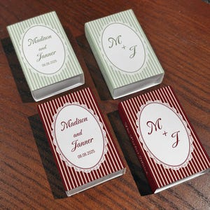 May include: Four rectangular matchboxes featuring vertical stripes in sage green or deep red. Each box displays a central white oval label with a lace-style border. The text reads Madison and Janner 08.08.2025 or the initials M + J.