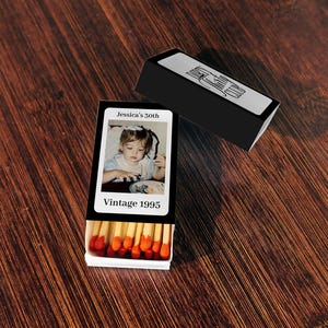 May include: A black matchbox with a photo of a young child and the text "Jessica's 30th" and "Vintage 1995." The matchbox is open, revealing red-tipped matches. A second black matchbox is also present.