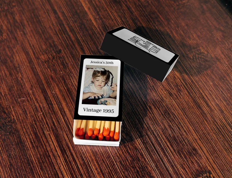 May include: A black matchbox with a photo of a young child and the text "Jessica's 30th" and "Vintage 1995." The matchbox is open, revealing red-tipped matches. A second black matchbox is also present.