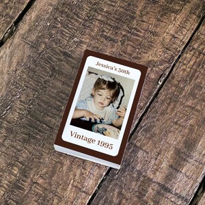 May include: A small, rectangular photo album with a brown cover. The album features a photograph of a young child and the text "Jessica's 50th" and "Vintage 1995". The album is resting on a wooden surface.