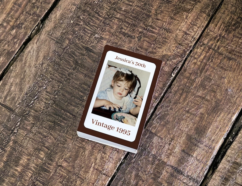 May include: A small, rectangular photo album with a brown cover. The album features a photograph of a young child and the text "Jessica's 50th" and "Vintage 1995". The album is resting on a wooden surface.