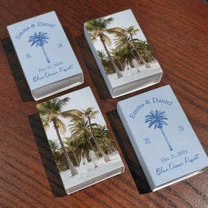 May include: Four light blue matchboxes with a palm tree design and the names "Emma & Daniel" printed in blue. The date "Dec 21, 2026" and "Blue Ocean Resort" are also printed on the matchboxes. The other sides of the matchboxes have a photo of palm trees.