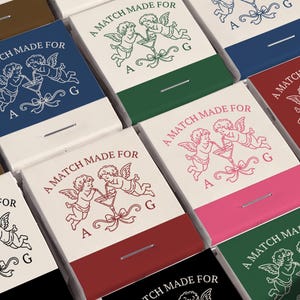May include: A collection of rectangular matchboxes featuring a graphic of two cherubs drinking from a martini glass with the text A MATCH MADE FOR A G. The boxes are available in various colors including blue, green, red, pink, and black.