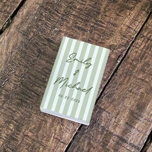 May include: A small, rectangular book with a light green and white striped cover. The cover features the names "Emily & Michael" in elegant script, along with the date "06.22.2026". The book rests on a textured, wooden surface.