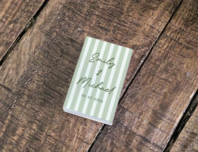 May include: A small, rectangular book with a light green and white striped cover. The cover features the names "Emily & Michael" in elegant script, along with the date "06.22.2026". The book rests on a textured, wooden surface.