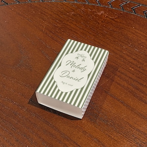 Peut inclure: Une bo&icirc;te d'allumettes rectangulaire avec un motif &agrave; rayures verticales vertes et blanches. Une &eacute;tiquette centrale blanche &agrave; bords festonn&eacute;s affiche le texte Melody & Daniel et la date Aug 6, 2026, avec un petit graphique de deux verres.