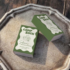 May include: Two green boxes with white text and graphics sit on a decorative plate. The boxes read "Camp Bach" and "Sabrina's Bachelorette Weekend, Lake Tahoe, California, 05.17.2026". The plate is a light brown color and has a textured surface.
