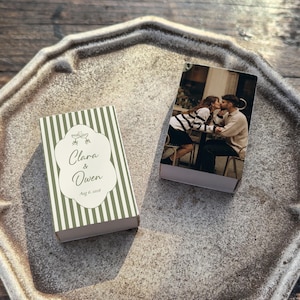 May include: Two small, rectangular boxes on a decorative tray. One box features a photo of a couple kissing. The other box has a green and white striped pattern with the names "Clara & Owen" and the date "Aug 6, 2016".