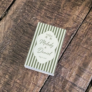 Peut inclure: Une bo&icirc;te d'allumettes rectangulaire avec un motif &agrave; rayures verticales vert sauge et blanc. Au centre, une &eacute;tiquette festonn&eacute;e blanche pr&eacute;sente un dessin de deux fl&ucirc;tes &agrave; champagne, les noms Melody & Daniel et la date Aug 8, 2026 en noir.
