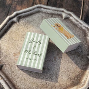 May include: Two small, light green striped boxes with the names "Emily & Michael" and the date "06.22.2026" printed on them. One box has a cartoon golden retriever wearing a yellow bandana. The boxes are on a decorative, octagonal plate.