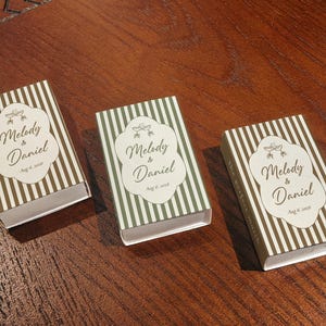 Peut inclure: Trois bo&icirc;tes d'allumettes rectangulaires orn&eacute;es de rayures verticales dans des tons marron et vert. Chaque bo&icirc;te pr&eacute;sente une &eacute;tiquette blanche centrale avec le texte Melody & Daniel, Aug 6, 2026, et deux coupes de champagne entrechoqu&eacute;es.