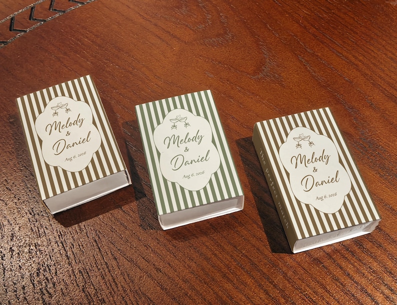 Peut inclure: Trois bo&icirc;tes d'allumettes rectangulaires orn&eacute;es de rayures verticales dans des tons marron et vert. Chaque bo&icirc;te pr&eacute;sente une &eacute;tiquette blanche centrale avec le texte Melody & Daniel, Aug 6, 2026, et deux coupes de champagne entrechoqu&eacute;es.