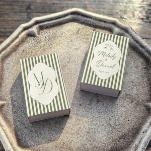 Peut inclure: Deux bo&icirc;tes d'allumettes rectangulaires avec un motif &agrave; rayures verticales vert olive et blanc. Une bo&icirc;te pr&eacute;sente le monogramme MD dans un cadre festonn&eacute; blanc, tandis que l'autre affiche le texte Melody & Daniel et Aug 16, 2026.