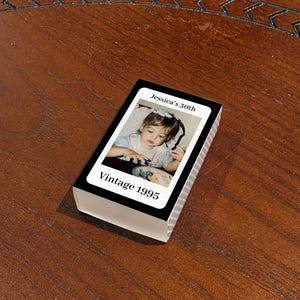 May include: A deck of playing cards with a photograph of a young child on the face of each card. The top card reads "Jessica's 50th" and "Vintage 1995" in white text on a black background. The deck is resting on a wooden surface.