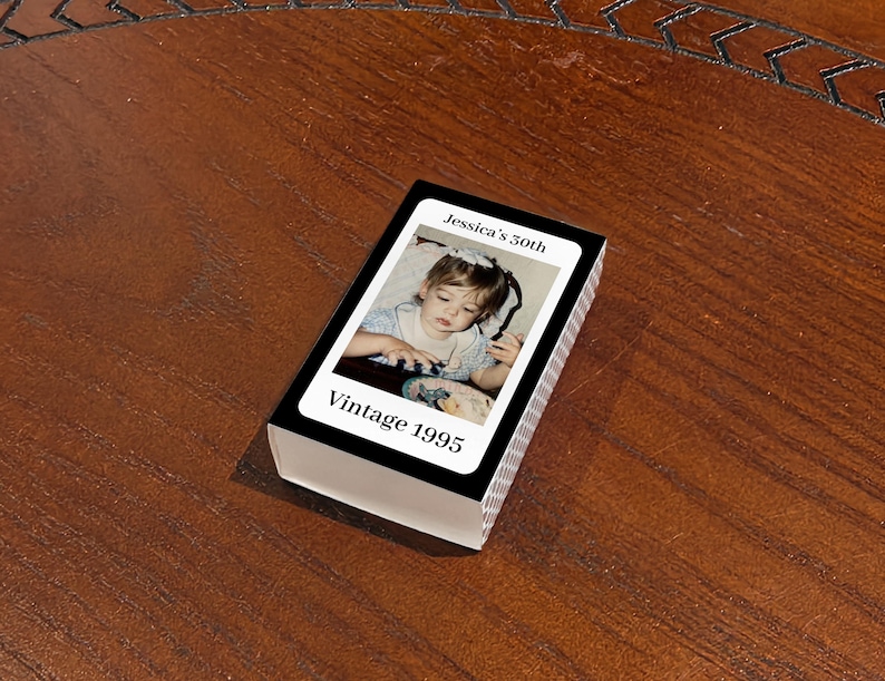 May include: A deck of playing cards with a photograph of a young child on the face of each card. The top card reads "Jessica's 50th" and "Vintage 1995" in white text on a black background. The deck is resting on a wooden surface.
