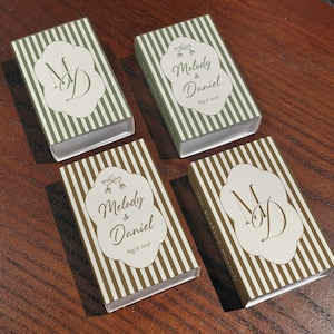 Peut inclure: Quatre bo&icirc;tes d'allumettes rectangulaires avec un motif &agrave; rayures verticales vert olive et blanc. Deux bo&icirc;tes pr&eacute;sentent le monogramme MD, tandis que deux autres portent le texte Melody & Daniel avec la date Aug 6, 2026 et deux coupes de champagne.