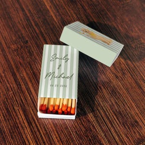May include: A matchbox with a light green and white striped design, featuring the names "Emily & Michael" and the date "06.22.2026". The box is open, revealing red-tipped matches. A second box with a dog illustration is also present.