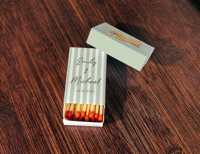 May include: A matchbox with a light green and white striped design, featuring the names "Emily & Michael" and the date "06.22.2026". The box is open, revealing red-tipped matches. A second box with a dog illustration is also present.