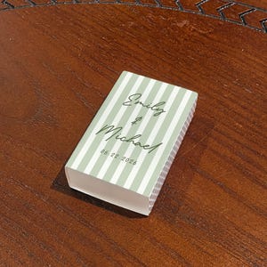 May include: A rectangular box with a light green and white striped pattern. The box has the names "Emily & Michael" and the date "06.22.2026" printed on the top. The box is resting on a dark brown wooden surface.