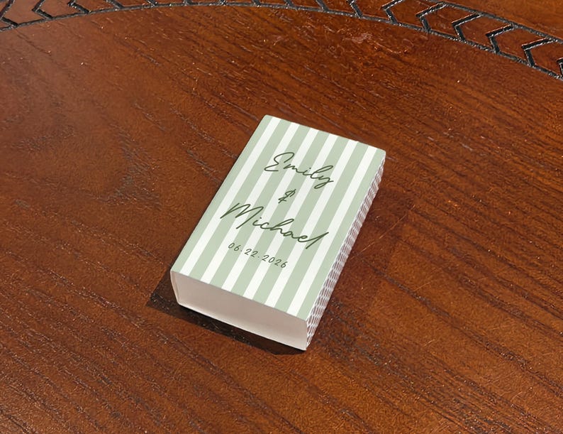 May include: A rectangular box with a light green and white striped pattern. The box has the names "Emily & Michael" and the date "06.22.2026" printed on the top. The box is resting on a dark brown wooden surface.