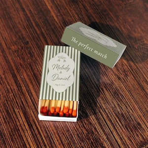 Peut inclure: Une bo&icirc;te d'allumettes rectangulaire &agrave; rayures verticales vert sauge et blanc. La bo&icirc;te pr&eacute;sente une &eacute;tiquette blanche avec le texte Melody & Daniel et Aug 8, 2026. Le couvercle coulissant affiche le texte The perfect match et un dessin de verres.